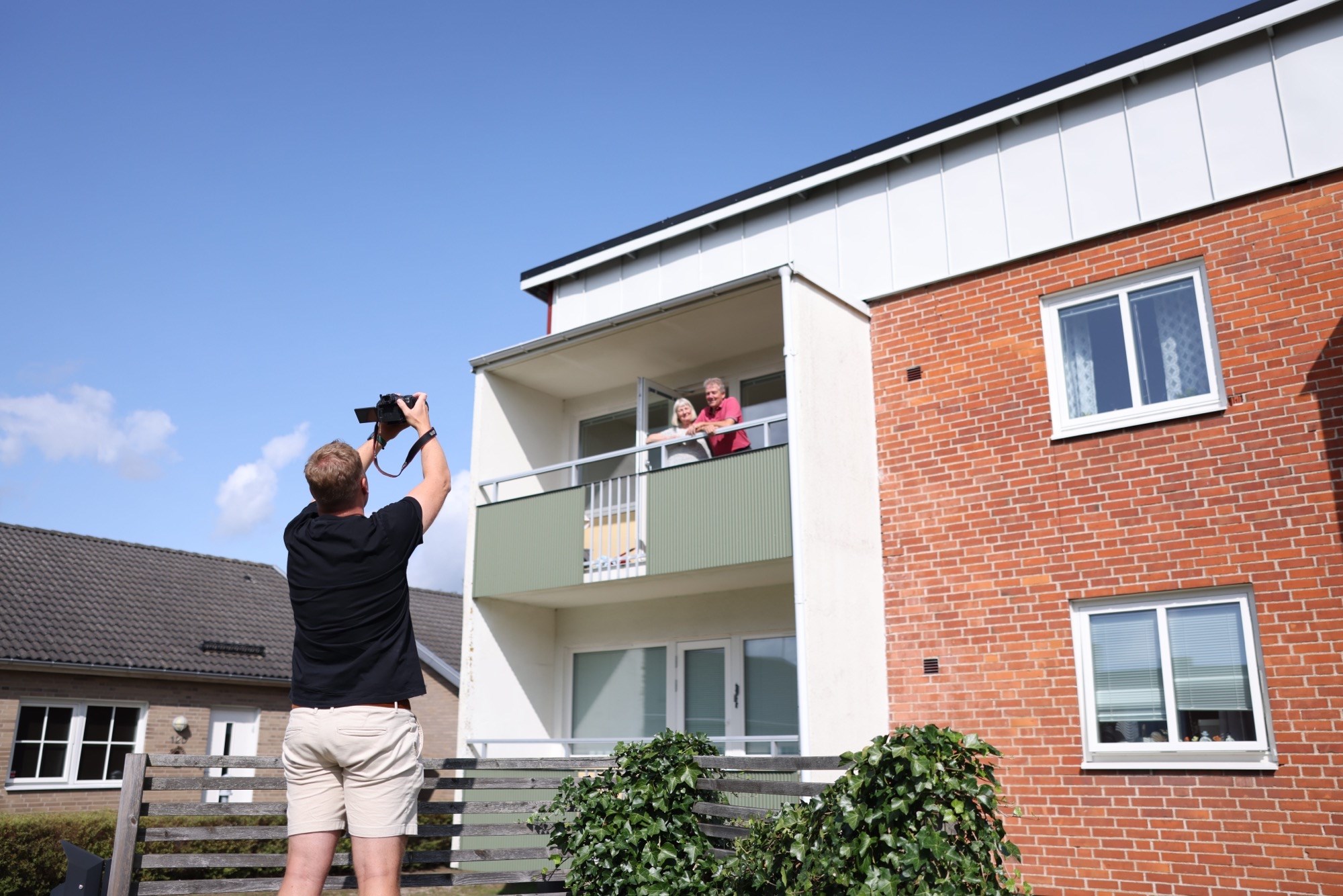 En kille står och fotograferar en man och en kvinna som står uppe på en balkong