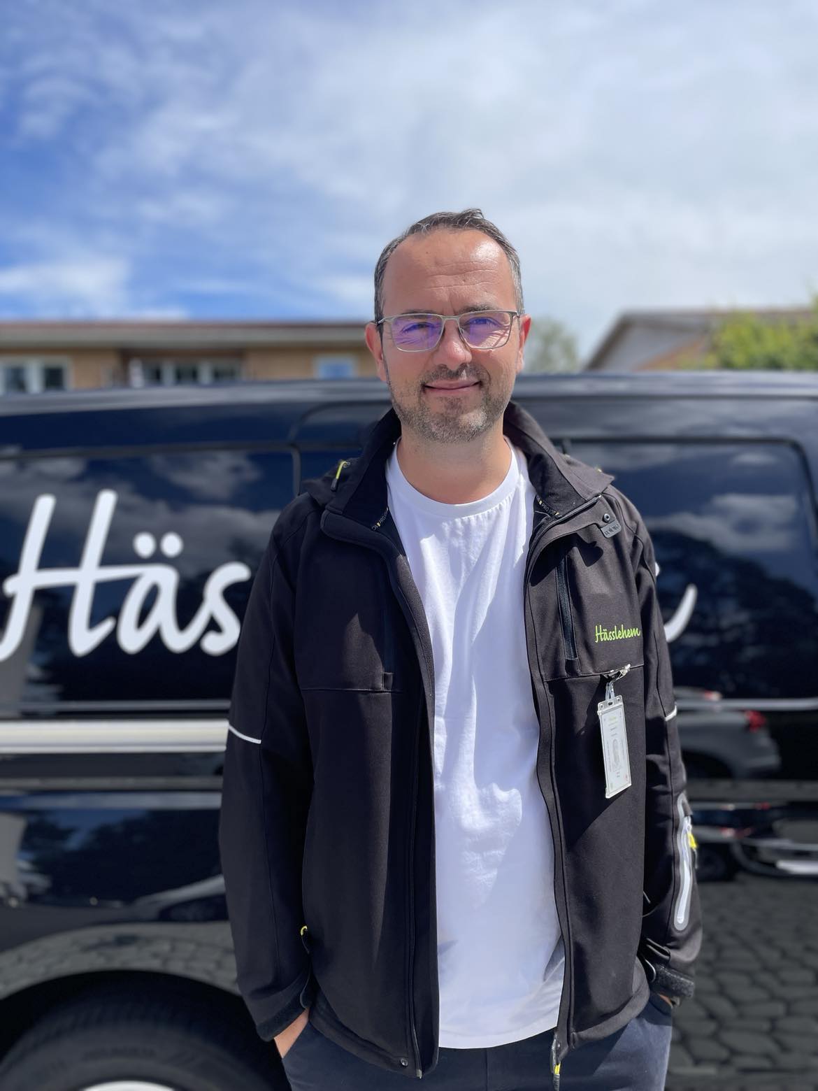 Man i svart jacka och vit tröja står framför en svart bil med Hässlehem logga