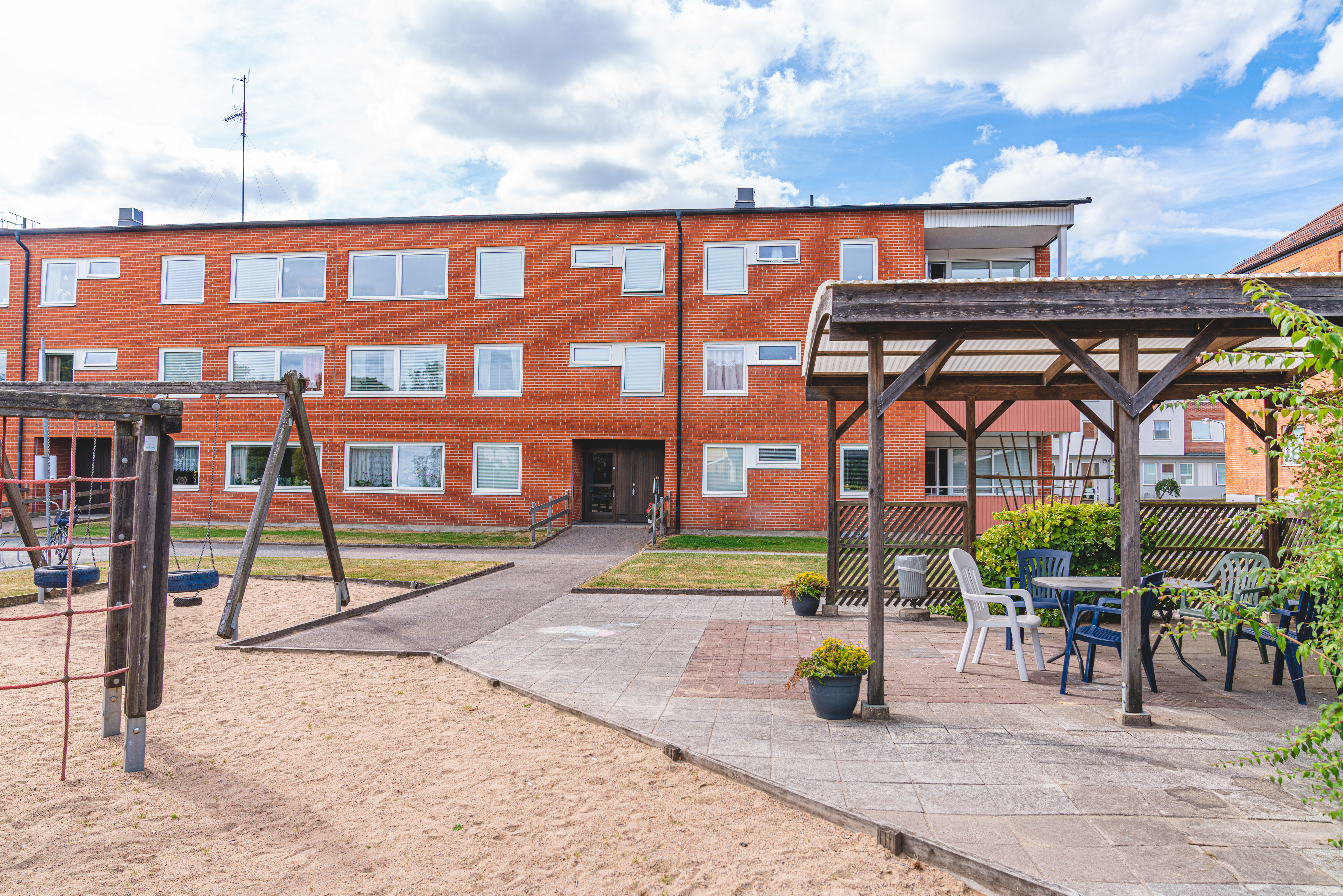 Trevåningshus i rött tegel med lekplats och pergola i förgrunden